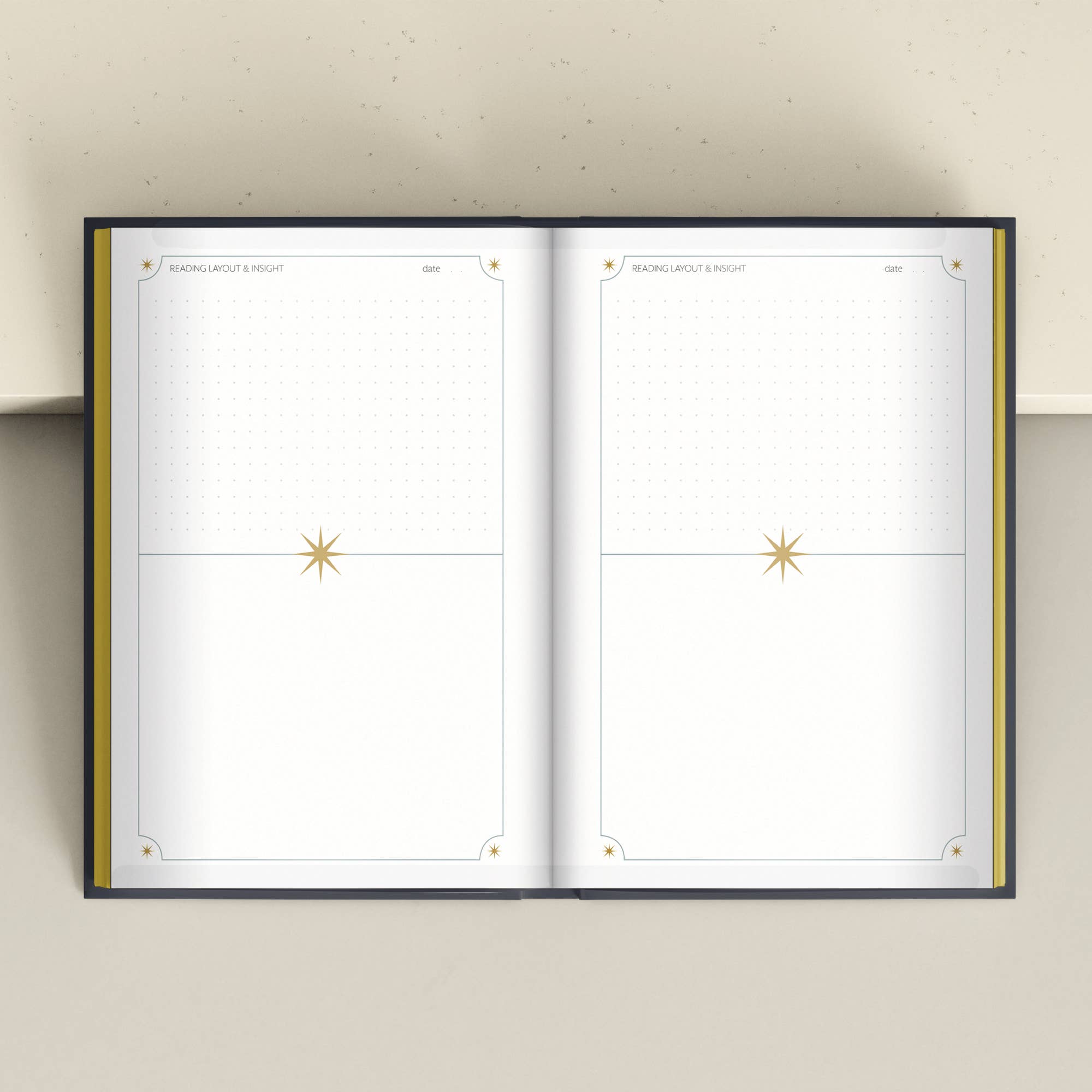 An open planner or notebook with two blank white interior spreads, each featuring a gold star motif near the center and subtle decorative corner details, photographed from above against a light beige background.