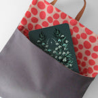 A dark gray tote bag with a red and pink polka-dot interior, holding a square card featuring a botanical illustration of white flowers on a dark green background.