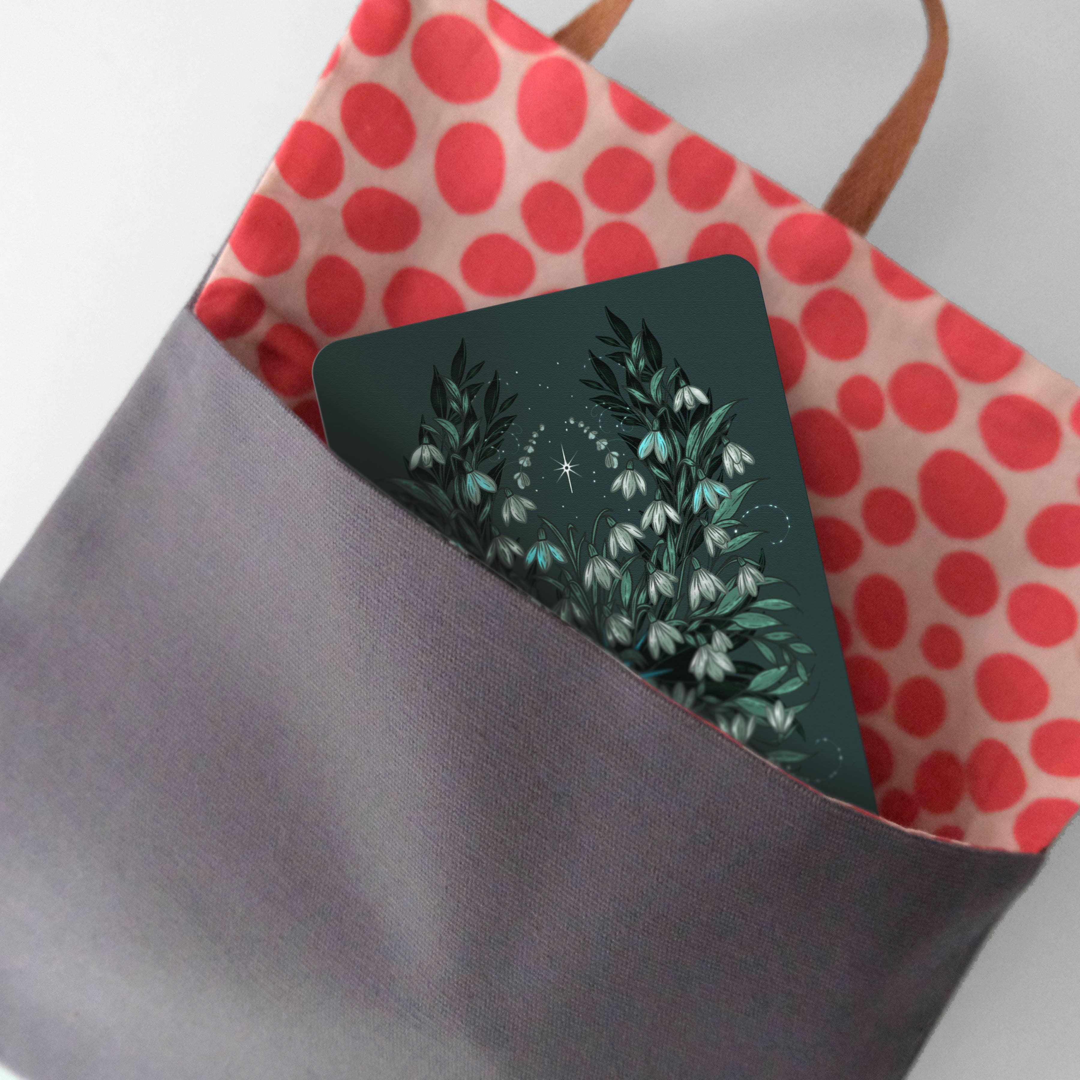 A dark gray tote bag with a red and pink polka-dot interior, holding a square card featuring a botanical illustration of white flowers on a dark green background.