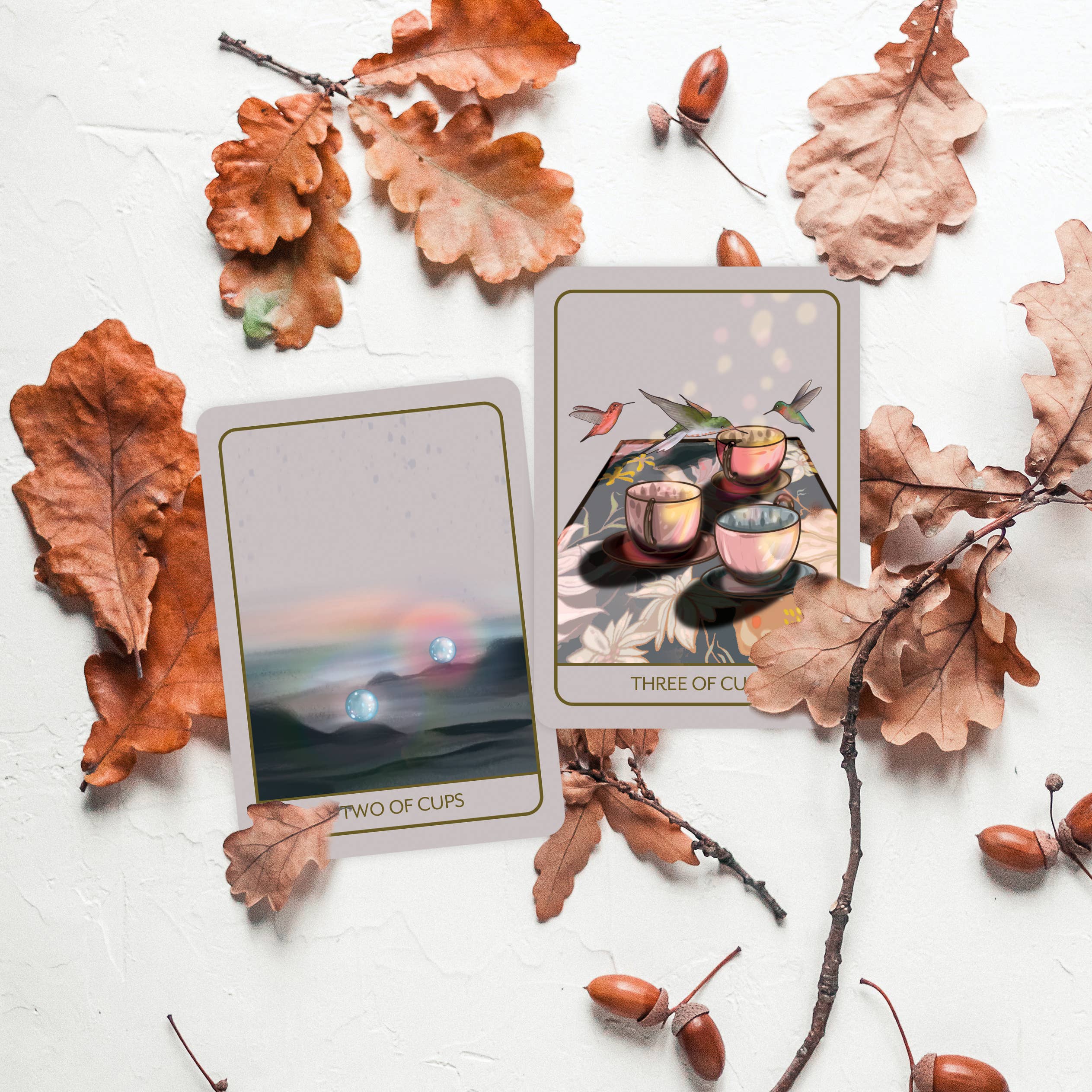 Two illustrated tarot cards, titled 'Two of Cups' and 'Three of Cups,' arranged on a white textured surface with scattered brown oak leaves and acorns.