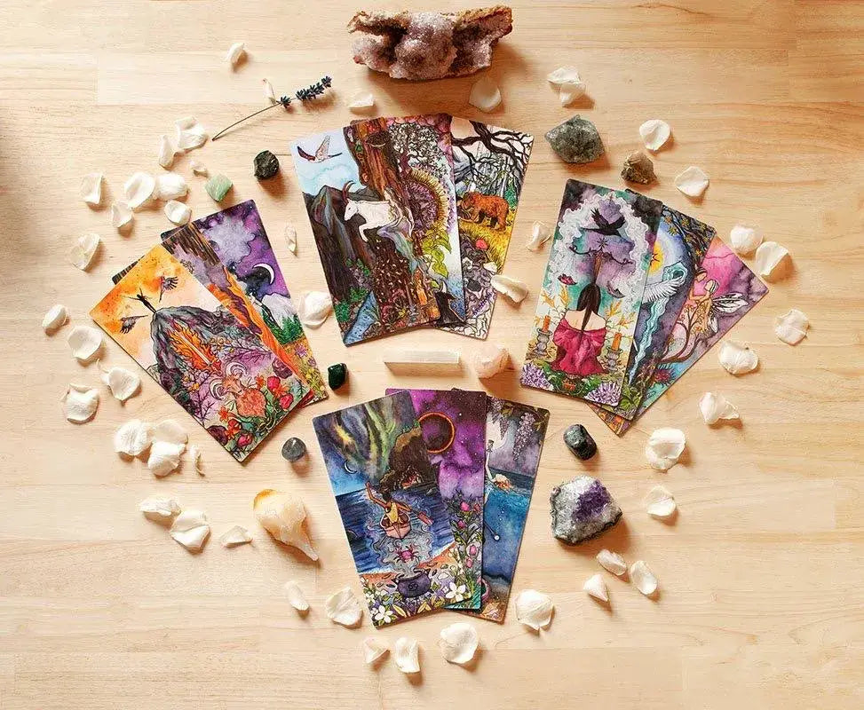 A set of illustrated tarot cards arranged in a circle on a light wood surface, surrounded by small white stones, crystals, and a few larger rocks. The cards feature colorful fantasy and nature scenes with figures, trees, and mystical imagery.