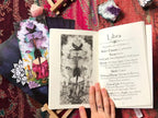 An open tarot deck booklet or journal lies on a patterned red textile, showing a black-and-white illustration on one page and a printed tarot page titled 'Libra' on the other. A hand with a ring holds the open pages.