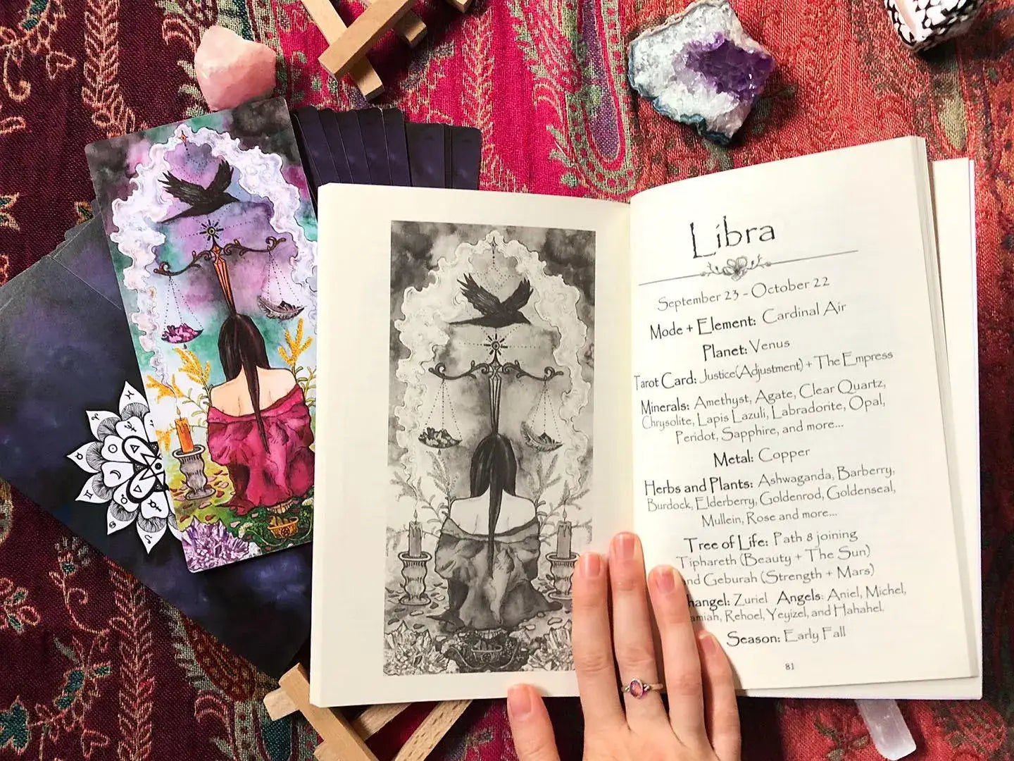 An open tarot deck booklet or journal lies on a patterned red textile, showing a black-and-white illustration on one page and a printed tarot page titled 'Libra' on the other. A hand with a ring holds the open pages.