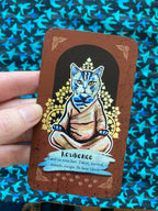 A hand holds a brown oracle card featuring an illustration of a meditating cat with the title 'Resilience' and a quote. The background is a blue patterned fabric.