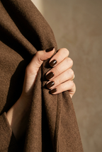 Hand holding brown fabric with dark nail polish against a neutral background