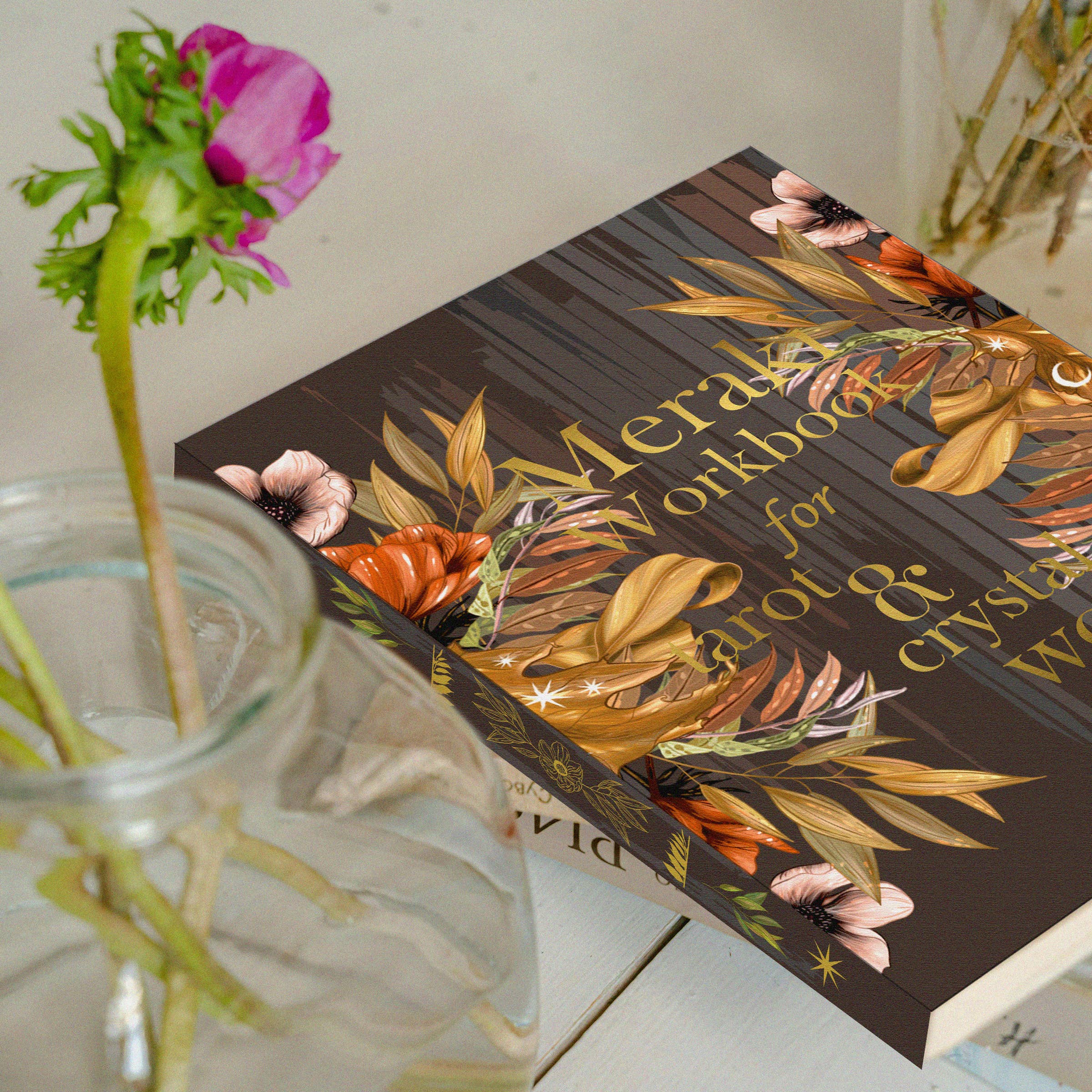 Decorative book with floral design on a surface next to a glass vase with flowers.