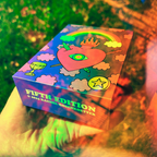 Colorful box with artistic design and 'Fifth Edition' text held in a hand against a blurred natural background.