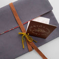 A close-up of a gray fabric-covered envelope or gift box tied with a brown woven ribbon and a small yellow bow, with a brown card reading 'Bouclier & Co' partially visible underneath on a light gray background.