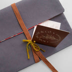 A close-up of a gray fabric-covered envelope or gift box tied with a brown woven ribbon and a small yellow bow, with a brown card reading 'Bouclier & Co' partially visible underneath on a light gray background.