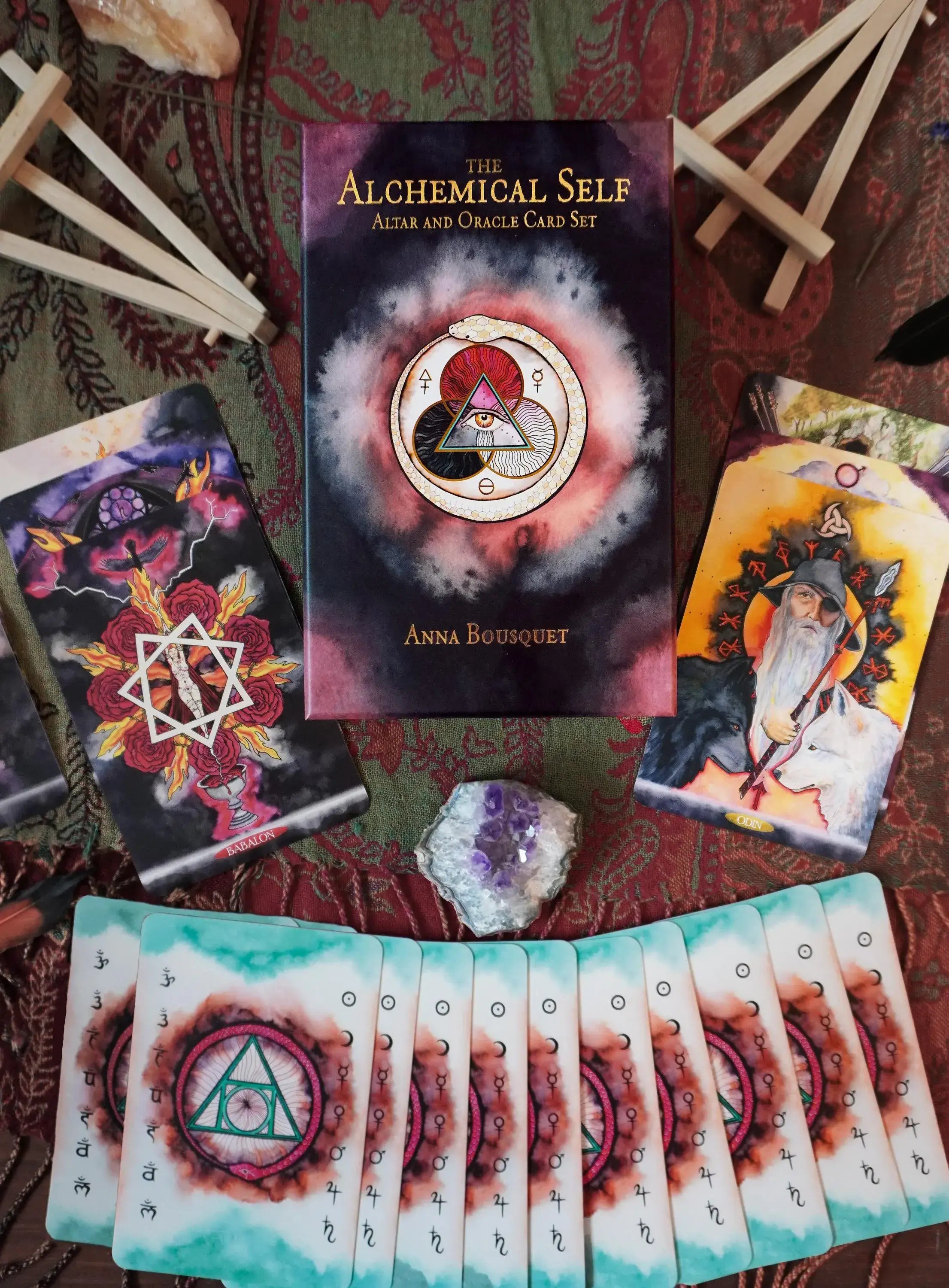 The Alchemical Self: Alka and Oracle Card Set by Anna Boussiquet, displayed in a styled flat-lay with tarot cards, crystals, and wooden sticks on a patterned red fabric background.