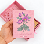 A hand holds a pink card featuring purple flowers and green leaves, with a matching pink box partially open behind it on a white background.