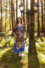 Woman standing in a forest with a lamp post, wearing a floral shawl and blue dress.
