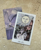 A tarot card deck featuring 'The Sun' card with a stylized face sun, horse, and figures, displayed on a textured beige stone surface next to a purple card.