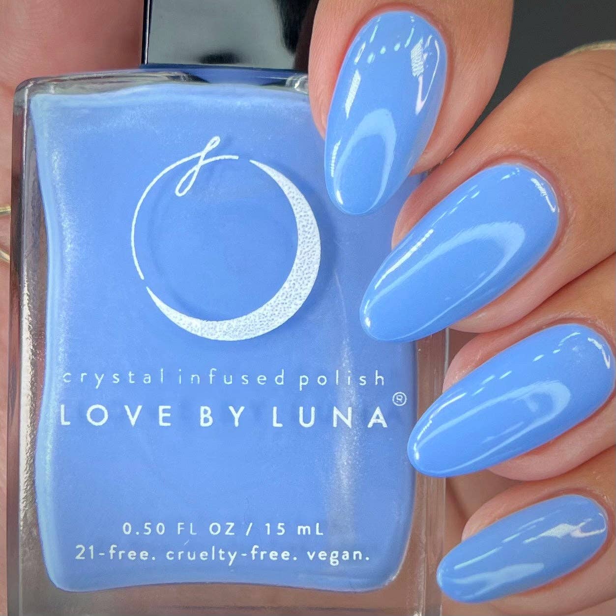 A close-up of a hand with long, glossy light blue manicured nails holding a bottle of Love By Luna crystal infused polish in a matching light blue shade.