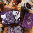 Purple tarot cards with black and white illustrations on a wooden surface with jars and dried flowers.