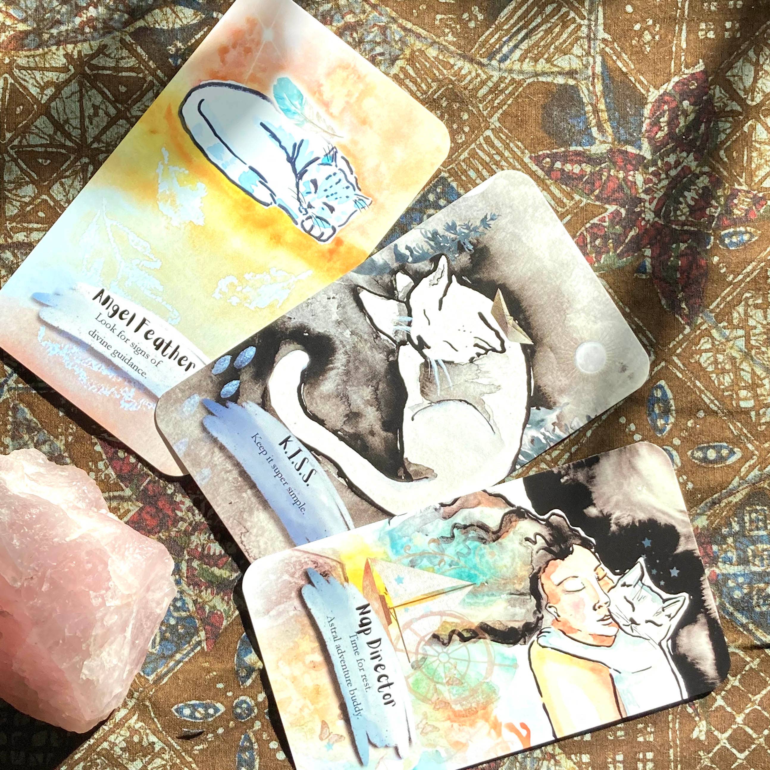 Three illustrated tarot cards featuring cats and a woman are arranged on a patterned brown fabric beside a pink crystal. The cards have watercolor-style artwork in soft pastel and dark ink tones, with visible text including 'My Beliefer' and 'K.