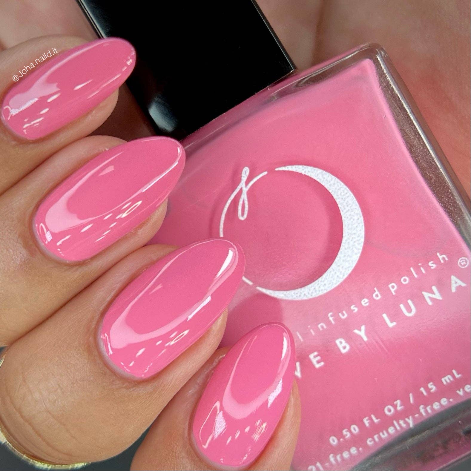 A close-up of a hand with glossy pink manicured nails holding a bottle of pink infused nail polish. The bottle label shows a white crescent moon logo and the brand name 'GIVE BY LUNA'.