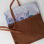 A brown fabric tote bag with a white printed floral panel and blue floral pattern, featuring brown woven handles, photographed on a light gray background.