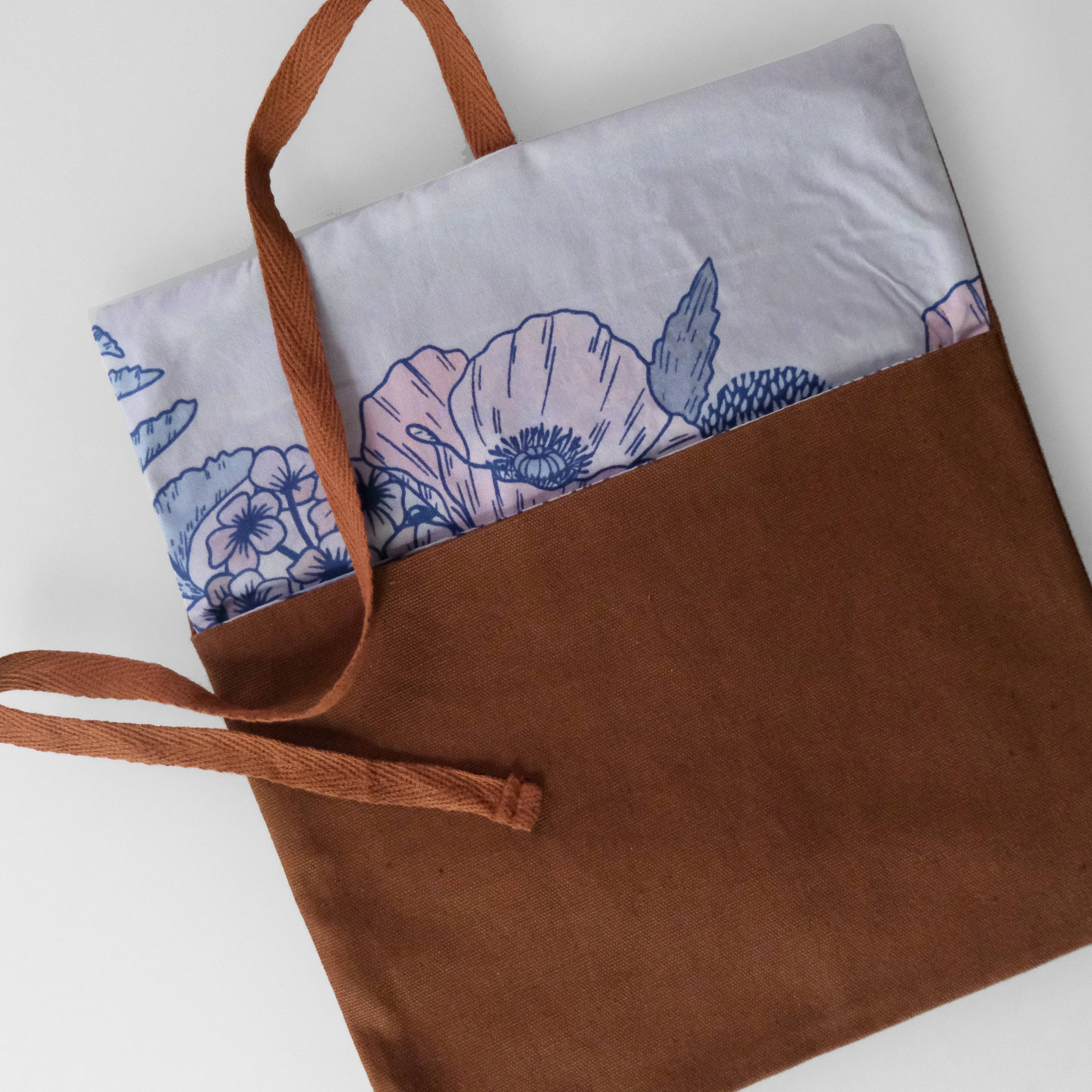 A brown fabric tote bag with a white printed floral panel and blue floral pattern, featuring brown woven handles, photographed on a light gray background.