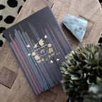 A dark blue-gray notebook with vertical wood-grain stripes and a gold foil 'Buckcherry Book Club' emblem, styled on a wooden table next to a crystal and a green plant.