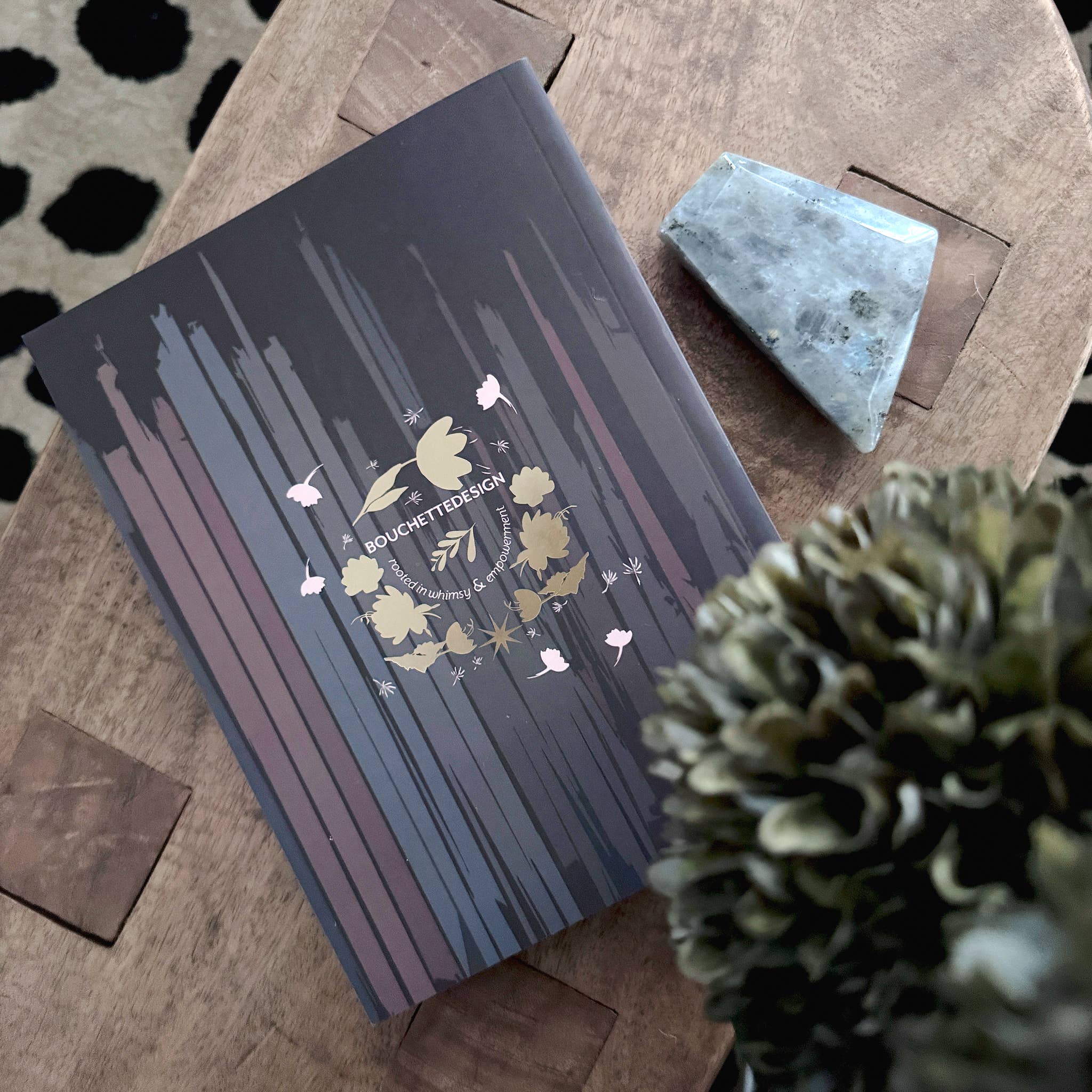 A dark blue-gray notebook with vertical wood-grain stripes and a gold foil 'Buckcherry Book Club' emblem, styled on a wooden table next to a crystal and a green plant.
