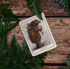 A tarot card titled 'Strength' featuring an illustration of a bear holding a pig, styled on a rustic wooden surface surrounded by green fern leaves.