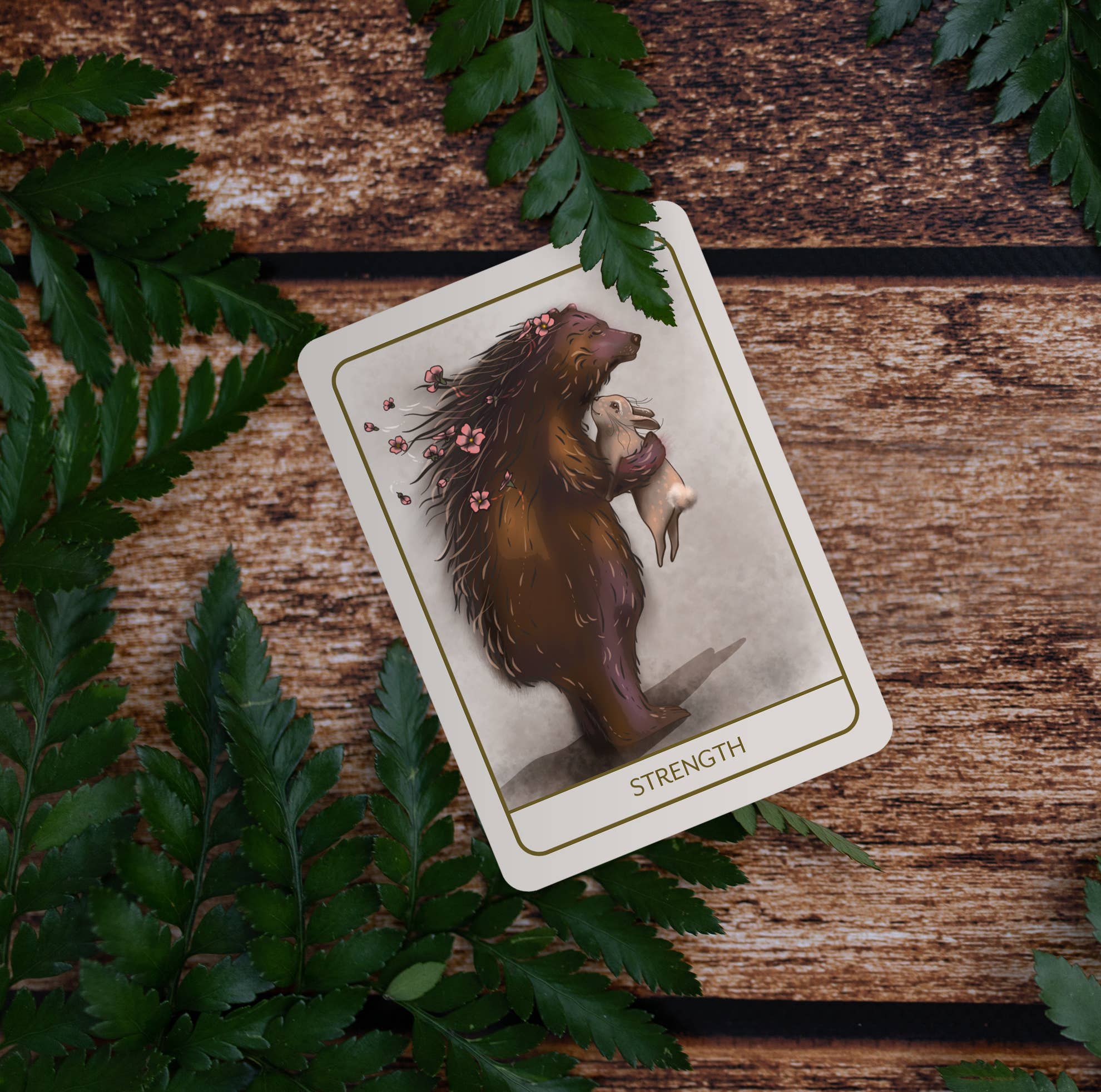 A tarot card titled 'Strength' featuring an illustration of a bear holding a pig, styled on a rustic wooden surface surrounded by green fern leaves.