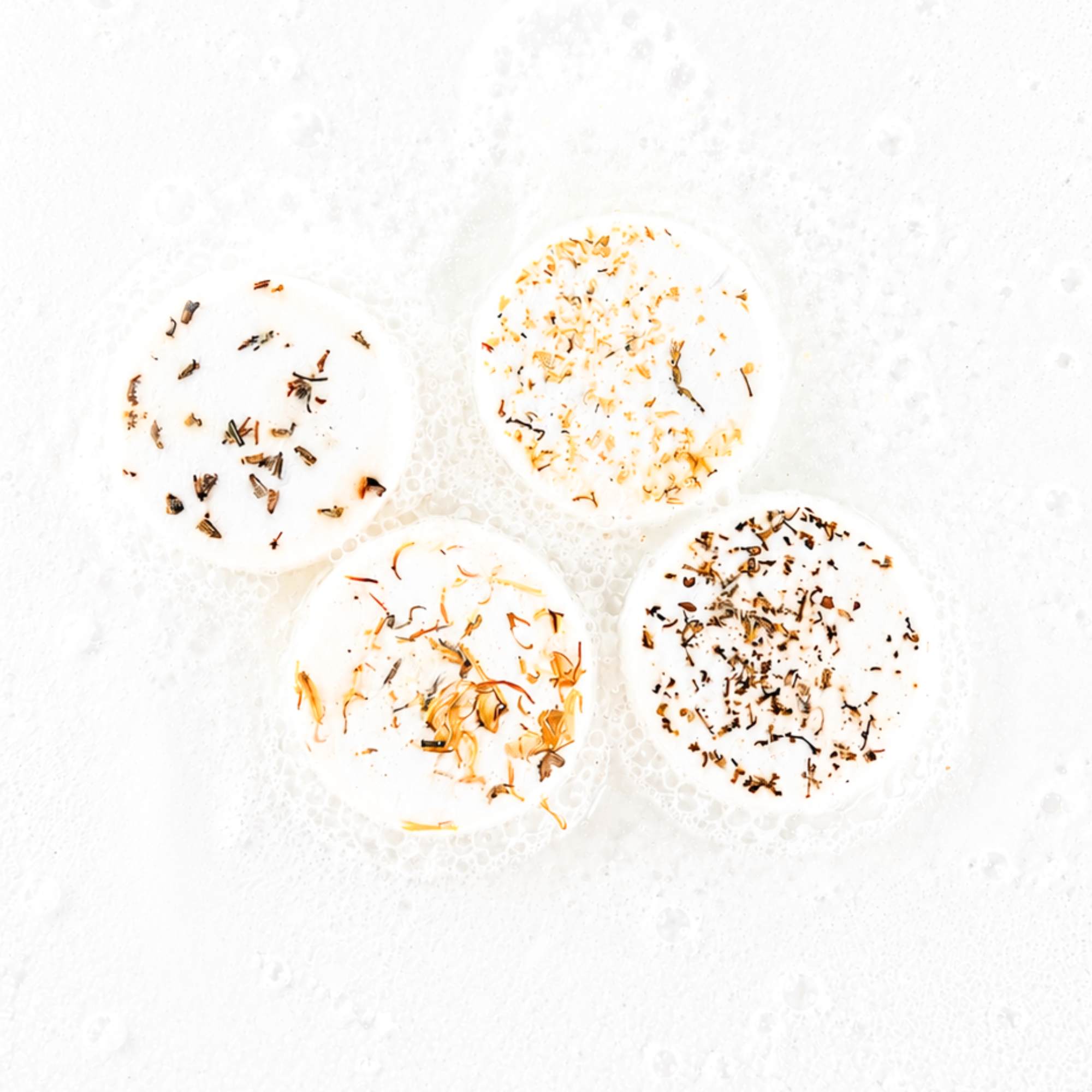 Four round bath bombs with different dried botanical toppings, arranged on a bubbly white surface.
