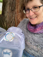 A smiling woman with glasses and a purple-and-pink knit scarf holds a clear plastic storage container wrapped in white tissue paper. The container has a round sticker with a blue silhouette of a dog and a circular label that reads 'Twin Lakes' with smaller text below.
