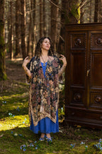 Woman wearing a patterned cardigan and blue dress standing in a forest with a wooden cabinet in the background.