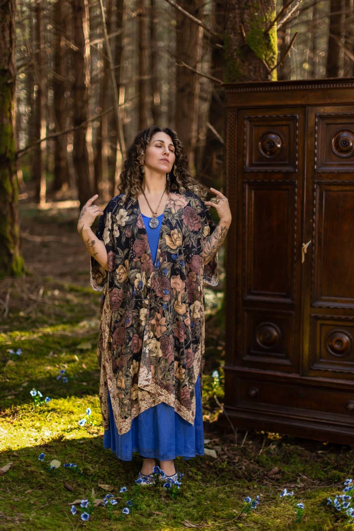 Woman wearing a patterned cardigan and blue dress standing in a forest with a wooden cabinet in the background.