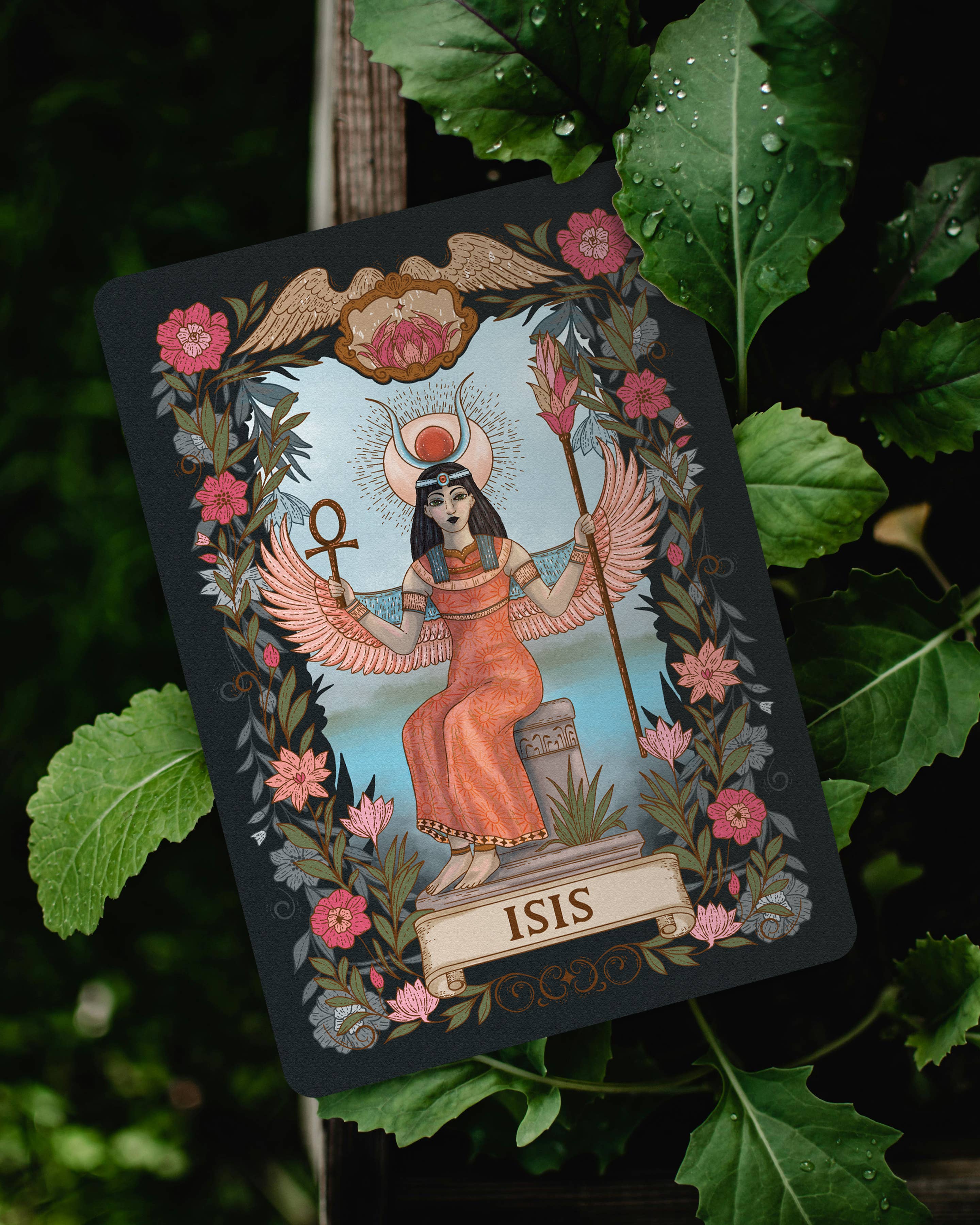 An illustrated tarot-style card featuring the goddess Isis, surrounded by pink flowers and green leaves, photographed outdoors against real foliage.