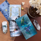 Set of tarot cards with artistic designs on a wooden surface with crystals and flowers.