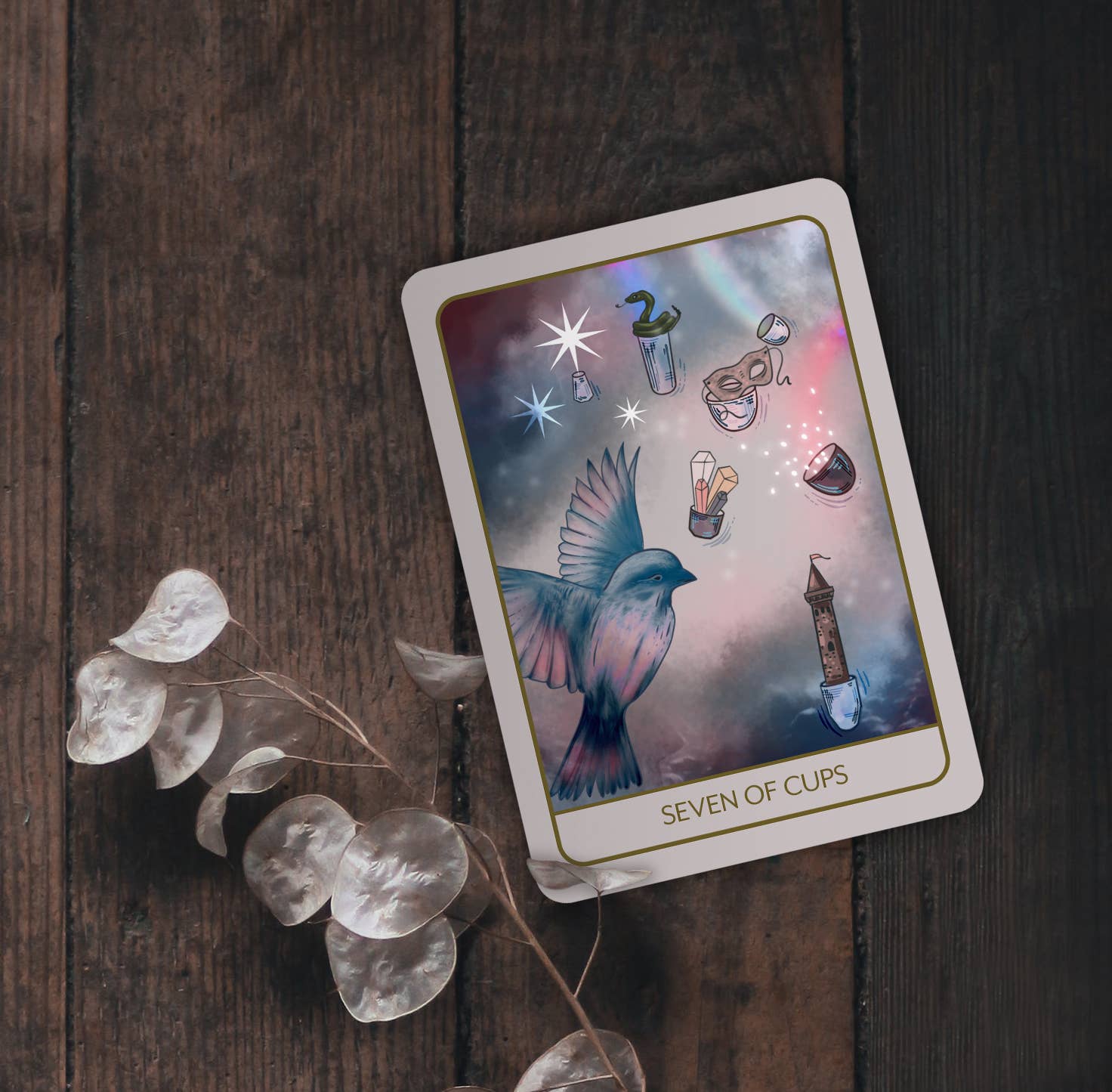 A tarot card titled 'SEVEN OF CUPS' lies on a dark wooden surface, styled with clear glass teardrop pendants and a sprig of dried leaves. The card features an illustration of a blue bird in flight against a pastel galaxy background with small icons of cups, glasses, stars, and a teapot.