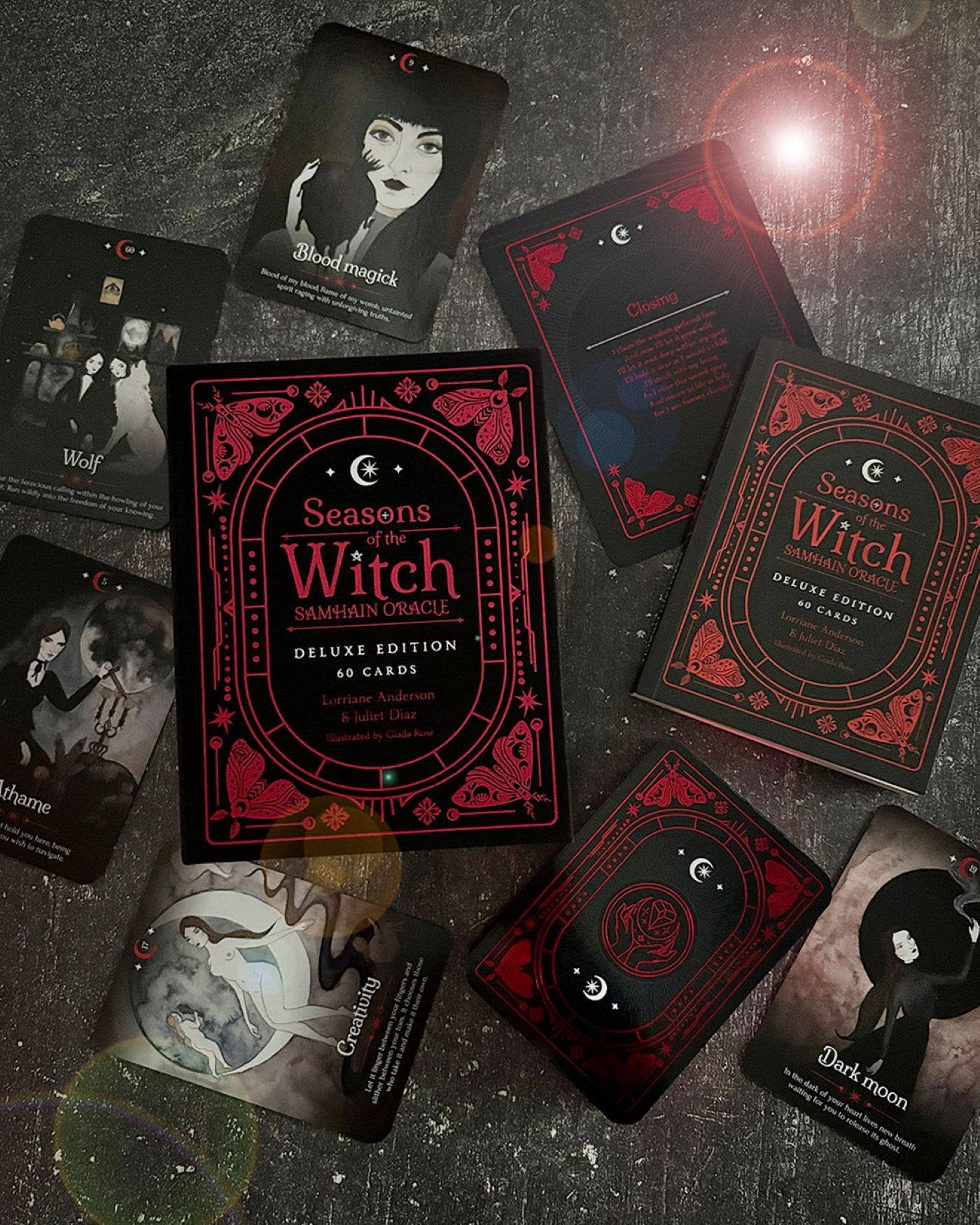 A flat-lay arrangement of the 'Seasons of the Witch Deluxe Edition' card deck and several illustrated cards on a dark textured surface. The packaging features a gothic red and black design with ornate borders, and the cards display dark, moody witch-themed illustrations.