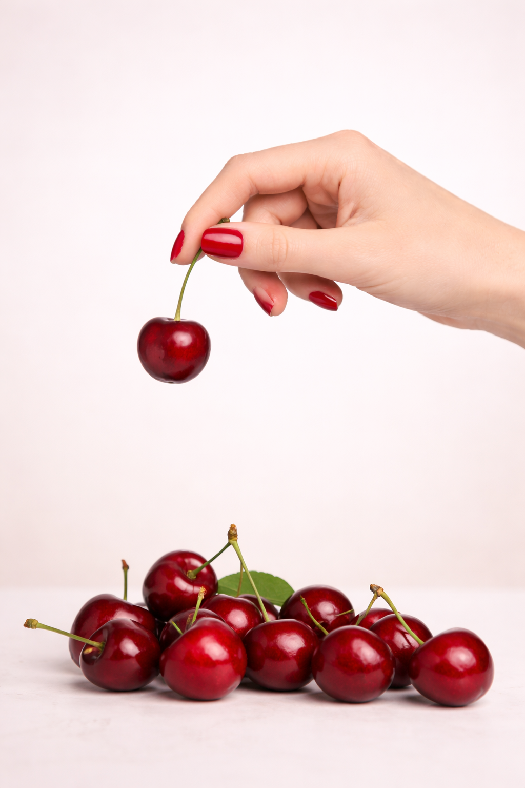 Hand holding a cherry with a pile of cherries on a light pink background