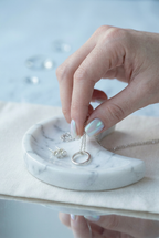 Hand holding a necklace over a marble jewelry dish with rings on a light blue background