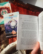 A hand holding an open book titled 'The Selkie's Oracle' while another book or booklet lies open beside it, showing illustrated oracle card artwork with the number 23 and a tarot-style card. The scene includes other books and printed materials on a tabletop.