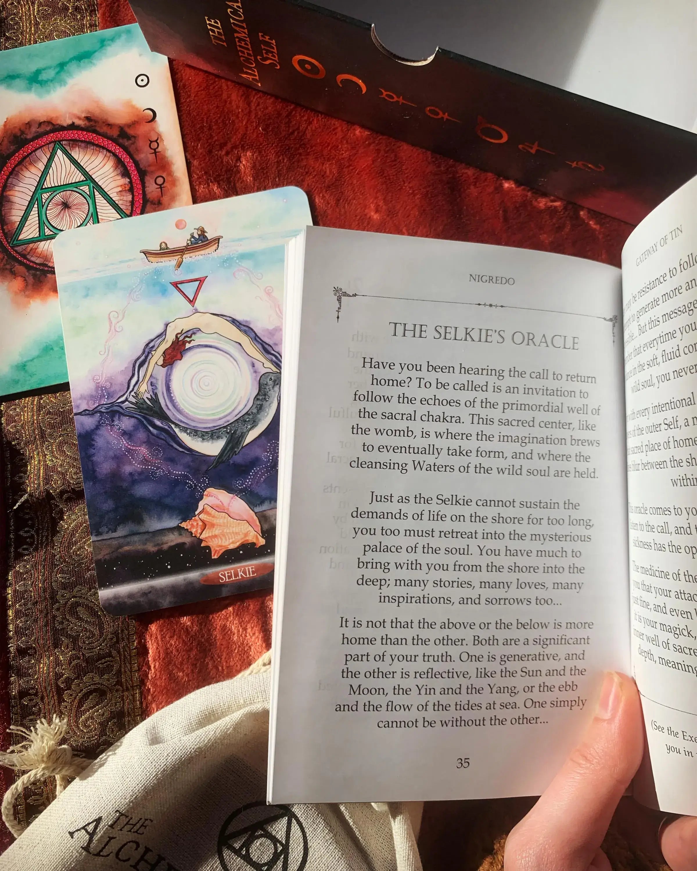 A hand holding an open book titled 'The Selkie's Oracle' while another book or booklet lies open beside it, showing illustrated oracle card artwork with the number 23 and a tarot-style card. The scene includes other books and printed materials on a tabletop.