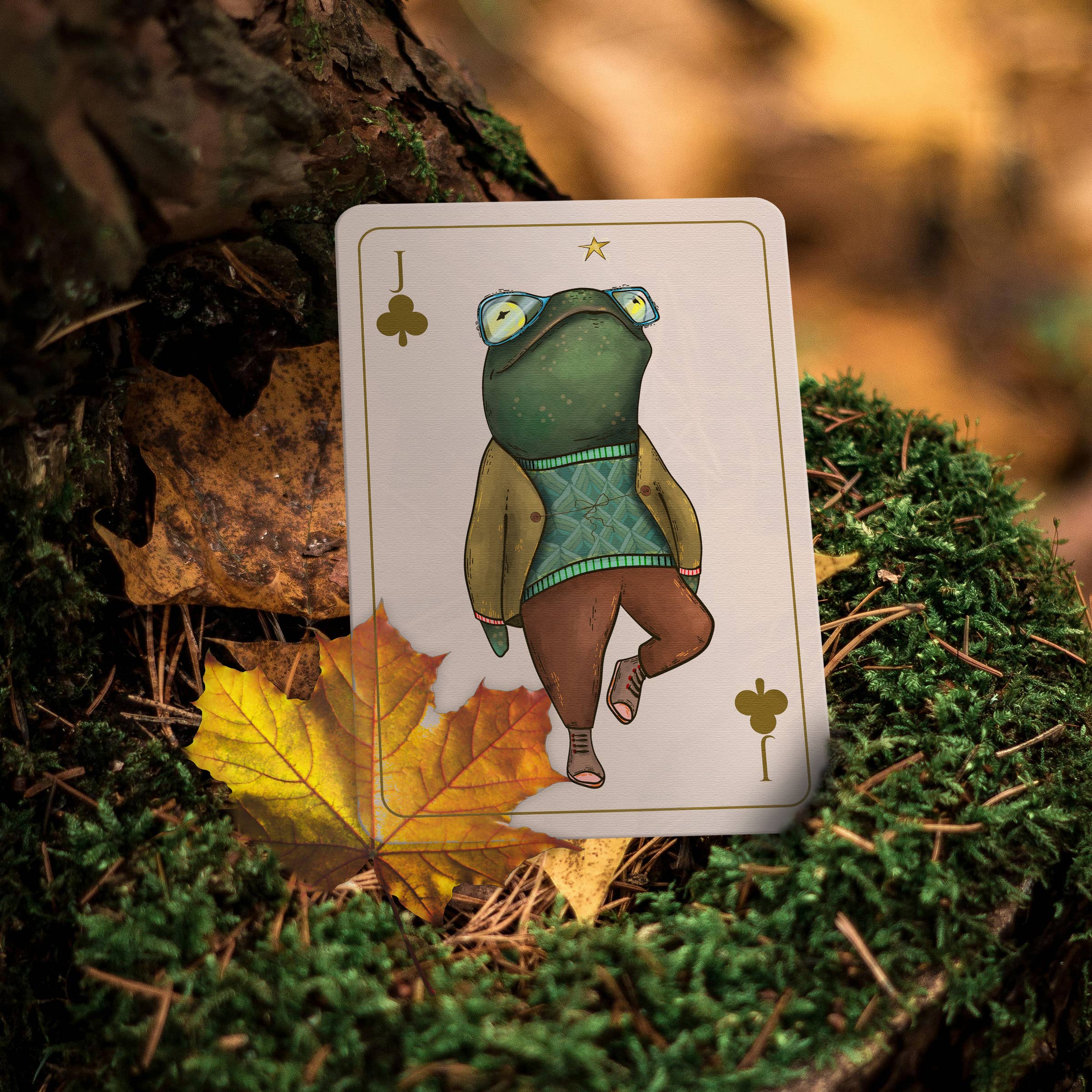 A playing card featuring an illustrated green frog wearing glasses, a tan jacket, and brown pants, placed on moss next to a yellow autumn leaf with a blurred forest background.