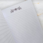 A close-up of a lined notepad page with a delicate floral line drawing at the top, photographed on a light marble-like surface.