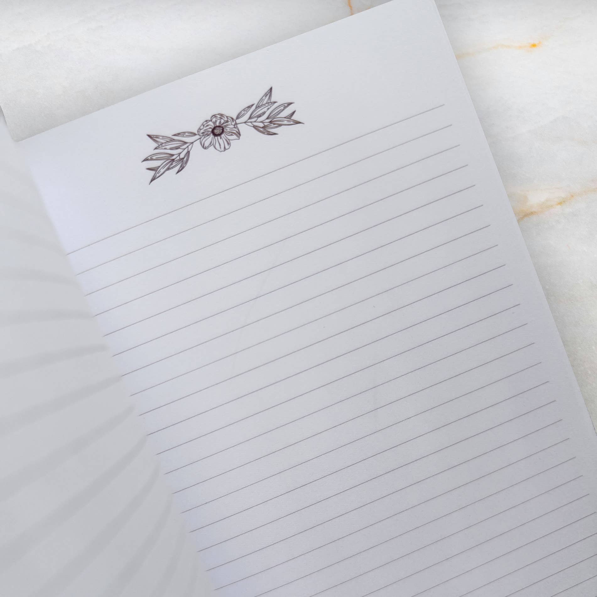 A close-up of a lined notepad page with a delicate floral line drawing at the top, photographed on a light marble-like surface.