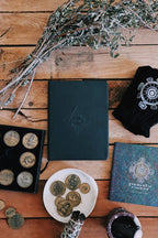 Dark green leather-bound book with embossed design on a wooden surface with coins, herbs, and a t-shirt.
