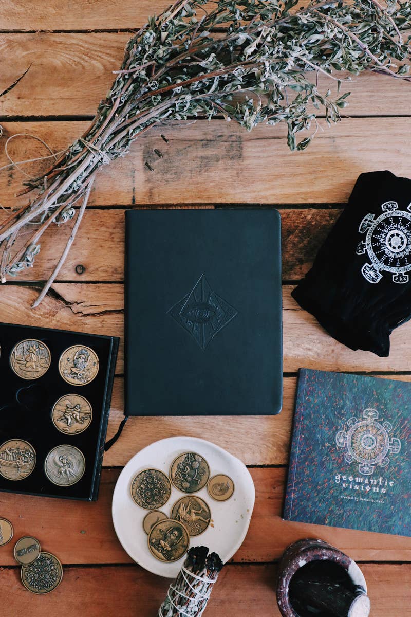 Dark green leather-bound book with embossed design on a wooden surface with coins, herbs, and a t-shirt.