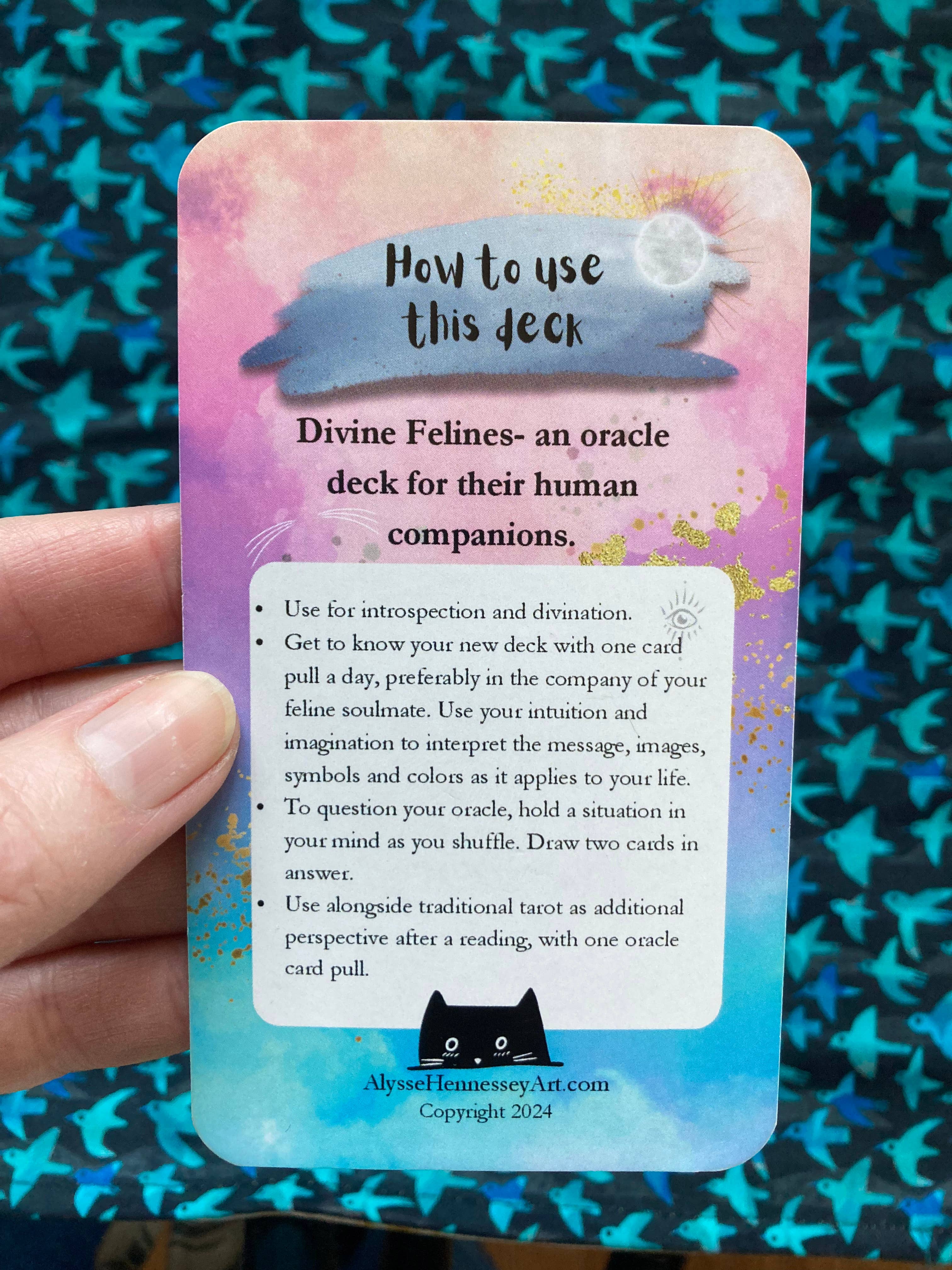 A hand holds an oracle card titled 'How to use this deck' for the 'Divine Felines- an oracle deck for their human companions' by Abyss Fairiesse's Art. The card features a pastel pink, purple, and blue gradient background with a black cat silhouette at the bottom, set against a blue patterned fabric backdrop.