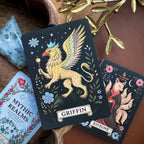 A styled flat-lay of a tarot deck featuring a 'Griffin' card with a golden winged lion and a 'Kitsune' card with a fox, surrounded by decorative props on a wooden surface.