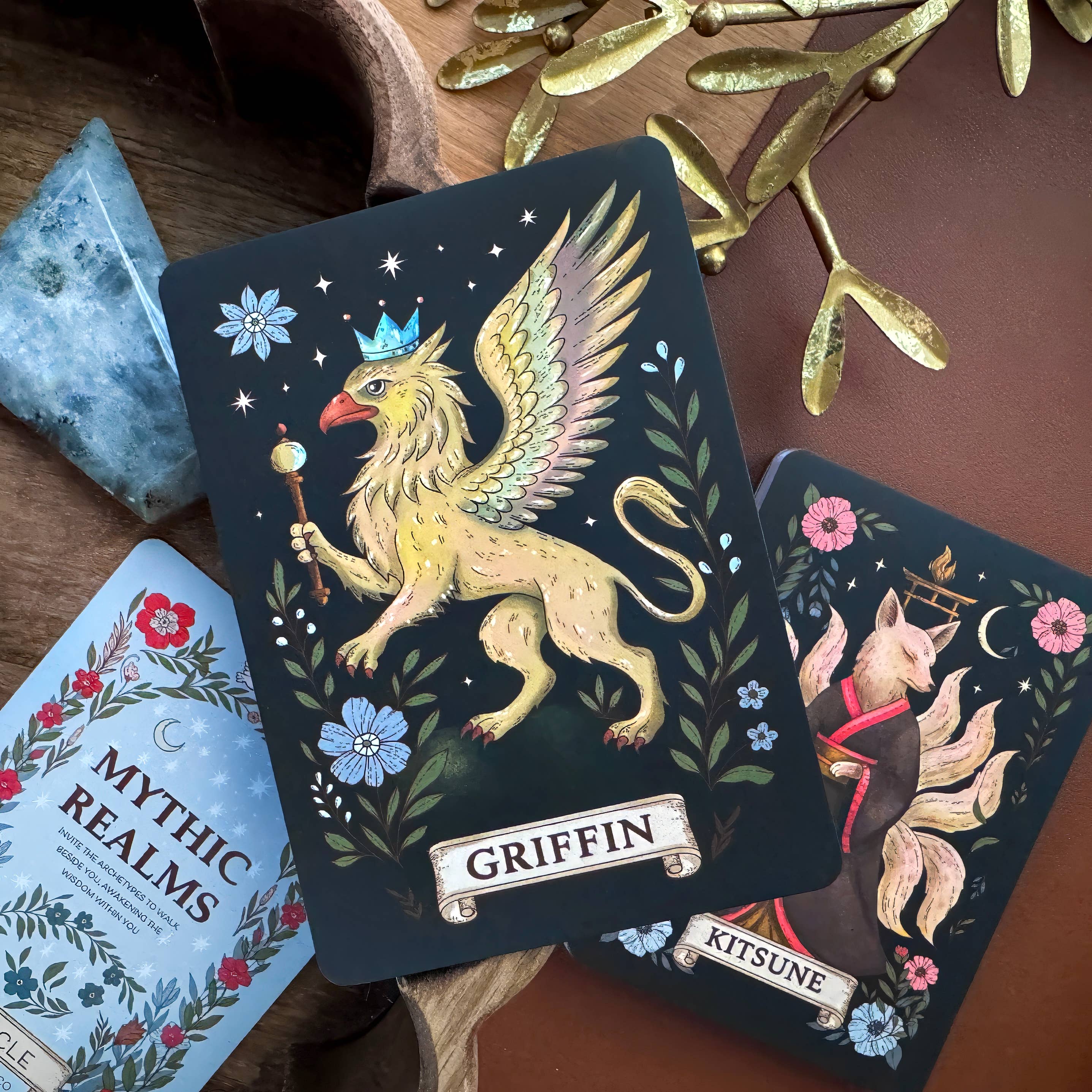 A styled flat-lay of a tarot deck featuring a 'Griffin' card with a golden winged lion and a 'Kitsune' card with a fox, surrounded by decorative props on a wooden surface.