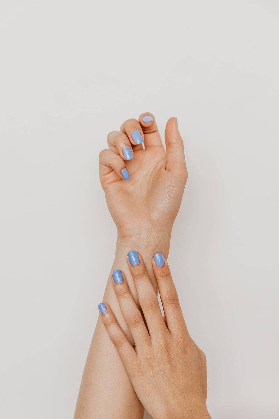 A close-up of two hands and forearms with neatly manicured fingernails painted in a light periwinkle blue shade, set against a clean, solid off-white background.