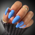 A close-up of a hand with long, almond-shaped nails painted in a glossy light blue color, wearing two gold rings and a dark gray knit sleeve against a dark gray background.