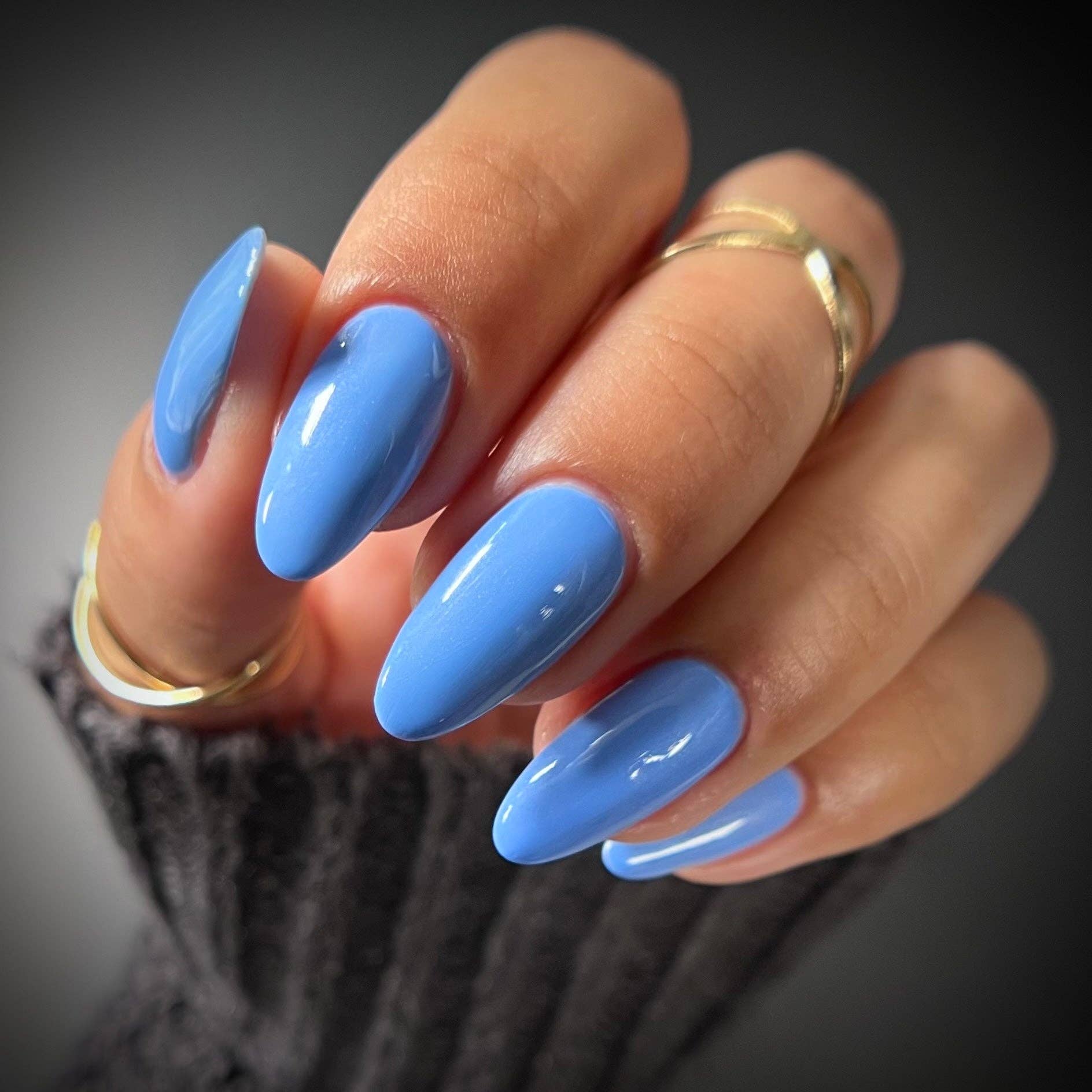 A close-up of a hand with long, almond-shaped nails painted in a glossy light blue color, wearing two gold rings and a dark gray knit sleeve against a dark gray background.